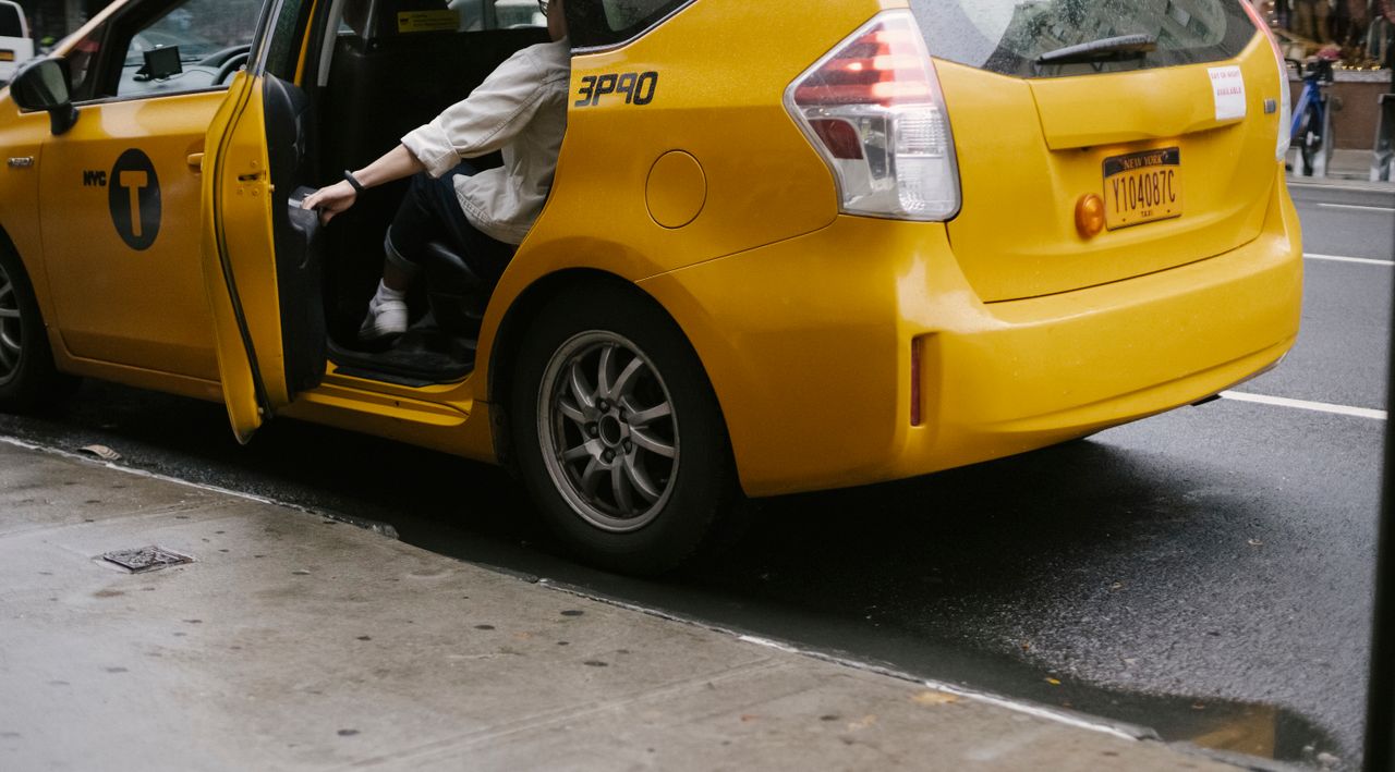 Unrecognizable passenger closing door of yellow cab with logo and headlight parked on asphalt road near pavement on street in city