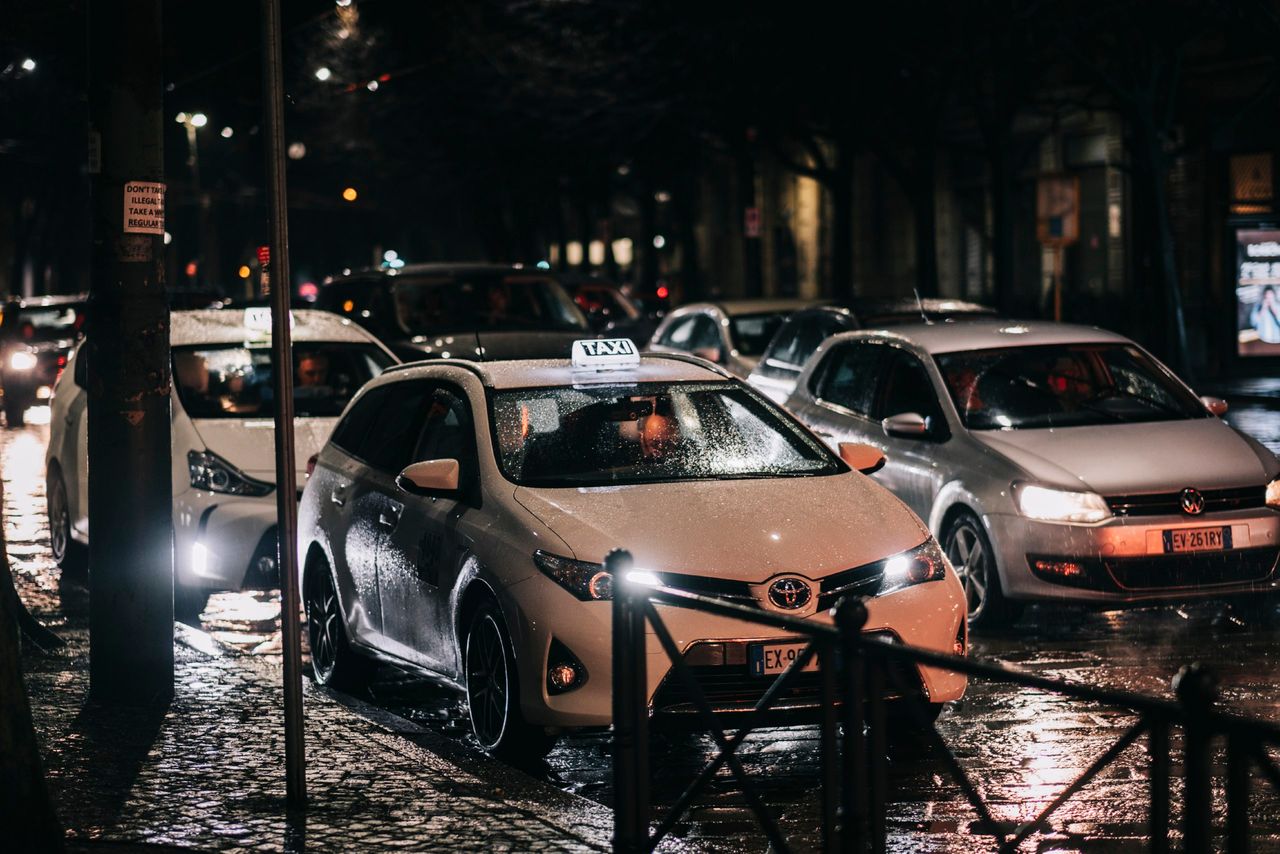 Busy city street at night with cars and taxis on a wet road, reflecting lights.